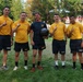 Five individuals wearing U.S. Navy physical training uniforms pose together on a grass field, with four of them holding up index fingers in a 'number one' gesture. The central figure holds a medicine ball or weighted exercise ball and wears a dark navy shirt with a military insignia, suggesting a leadership or instructor role. The setting appears to be an outdoor athletic or recreational event.