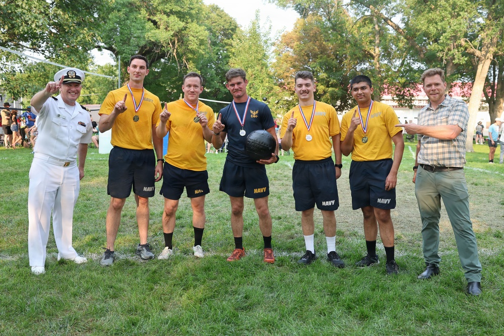A group of seven people pose for a victory photo on a grass field. Five individuals are wearing matching yellow 'NAVY' athletic uniforms with medals around their necks, suggesting they won a competition. They are flanked by a person in a white naval officer uniform on the left and a civilian in a plaid shirt on the right.