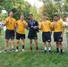 A group of seven people pose for a victory photo on a grass field. Five individuals are wearing matching yellow 'NAVY' athletic uniforms with medals around their necks, suggesting they won a competition. They are flanked by a person in a white naval officer uniform on the left and a civilian in a plaid shirt on the right.