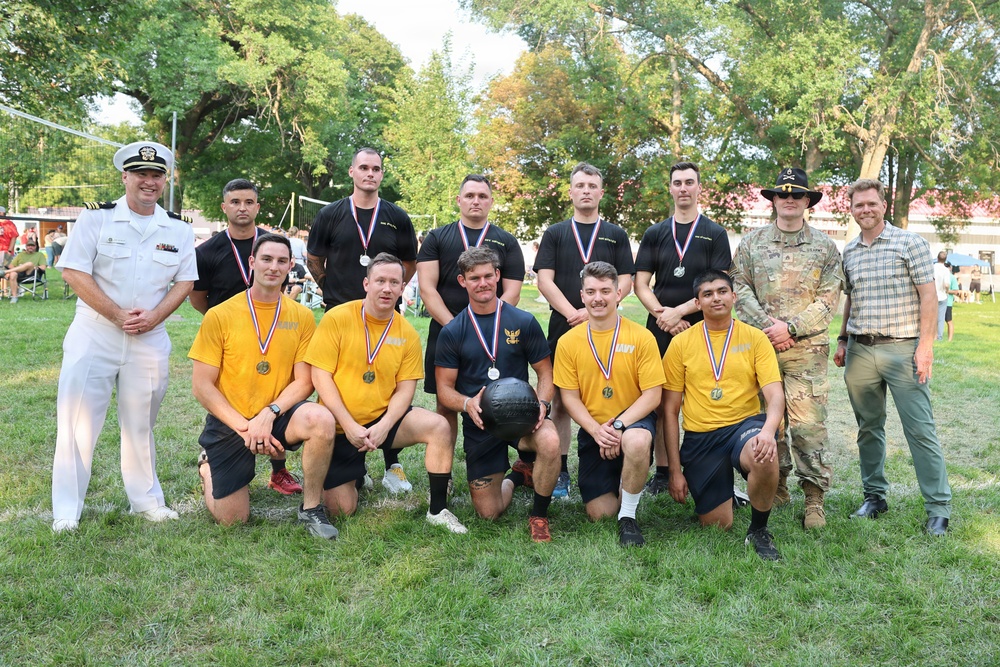 A group photo of military personnel posing outdoors on a grass field, likely after a sporting competition. The group includes members wearing Navy gold shirts, others in black athletic shirts, a Navy officer in white dress uniform, an Army soldier in camouflage, and a civilian. Several individuals are wearing medals around their necks, and one person is holding a medicine ball.