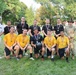 A group photo of military personnel posing outdoors on a grass field, likely after a sporting competition. The group includes members wearing Navy gold shirts, others in black athletic shirts, a Navy officer in white dress uniform, an Army soldier in camouflage, and a civilian. Several individuals are wearing medals around their necks, and one person is holding a medicine ball.