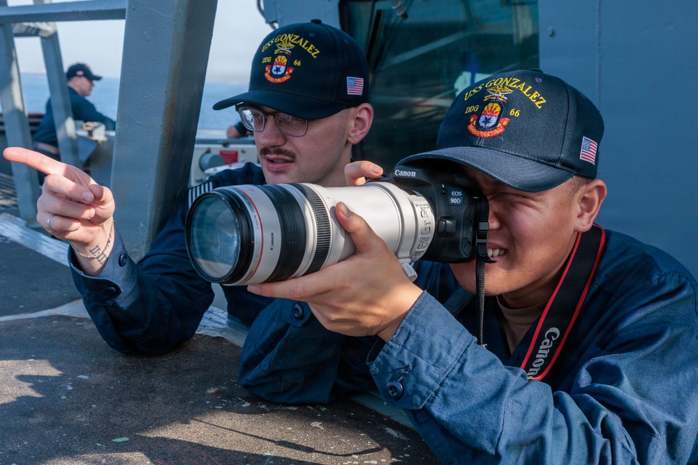 Two U.S. Navy sailors aboard the USS Gonzalez (DDG-66) are engaged in photography on the ship's deck. One sailor is looking through a Canon EOS 90D camera fitted with a large white Canon telephoto lens, while the other points in a direction, likely guiding the shot. A third crew member is visible seated in the background.