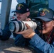 Two U.S. Navy sailors aboard the USS Gonzalez (DDG-66) are engaged in photography on the ship's deck. One sailor is looking through a Canon EOS 90D camera fitted with a large white Canon telephoto lens, while the other points in a direction, likely guiding the shot. A third crew member is visible seated in the background.