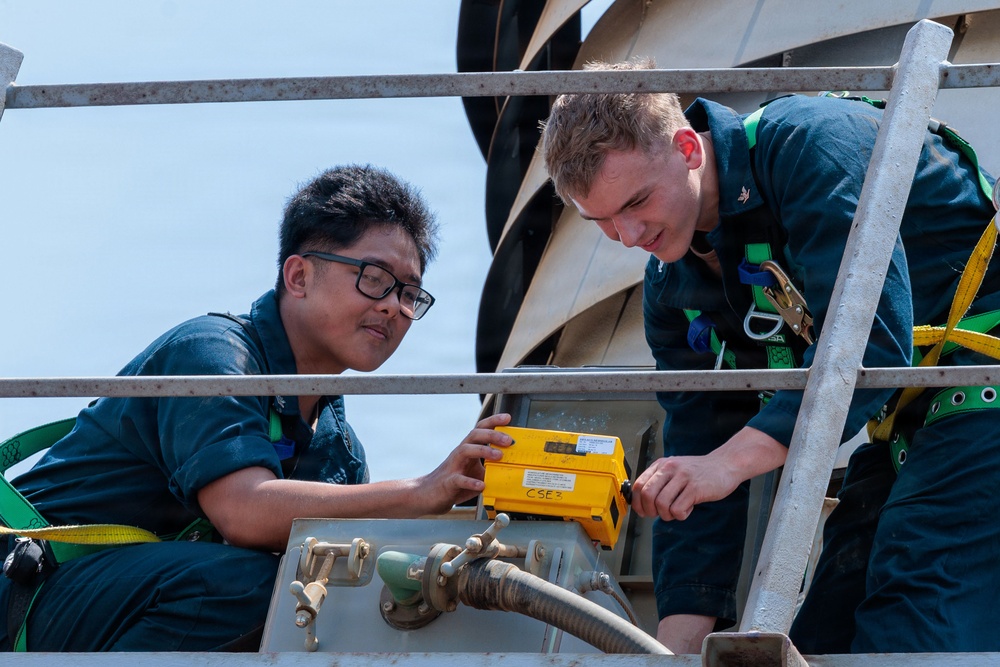 Two personnel in dark blue uniforms and safety harnesses are working together on what appears to be a naval vessel or industrial platform. They are examining or operating a yellow electronic device labeled 'CSE3' mounted on a metal surface. The setting appears to be an elevated outdoor work area with metal railings and piping visible.