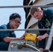 Two personnel in dark blue uniforms and safety harnesses are working together on what appears to be a naval vessel or industrial platform. They are examining or operating a yellow electronic device labeled 'CSE3' mounted on a metal surface. The setting appears to be an elevated outdoor work area with metal railings and piping visible.