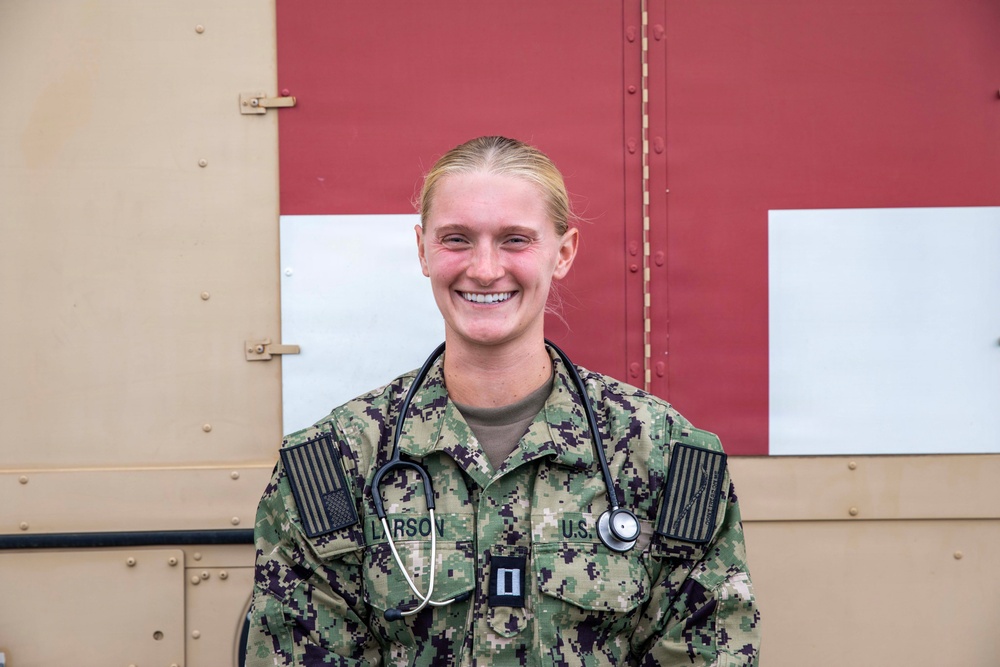 A smiling female military medical officer wearing U.S. Navy Working Uniform (NWU Type III) with a stethoscope around her neck. Her name tape reads 'LARSON' and she displays a Captain's rank insignia (O-3) on her chest. She is posed in front of what appears to be a military medical facility or vehicle with a red cross marking.