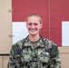 A smiling female military medical officer wearing U.S. Navy Working Uniform (NWU Type III) with a stethoscope around her neck. Her name tape reads 'LARSON' and she displays a Captain's rank insignia (O-3) on her chest. She is posed in front of what appears to be a military medical facility or vehicle with a red cross marking.