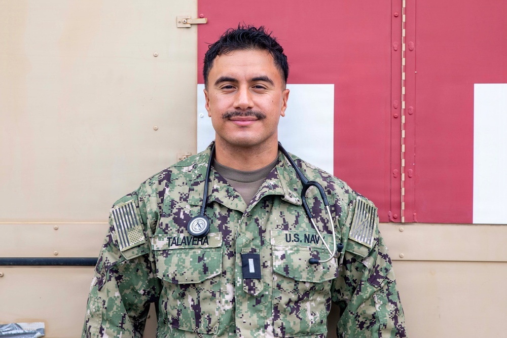 A U.S. Navy service member named Talavera is photographed in a portrait-style image wearing Navy Working Uniform (NWU) camouflage. The individual has a stethoscope around their neck and a rank insignia on their chest, indicating a medical officer role. They are standing in front of what appears to be a military medical vehicle or container marked with a red cross symbol.