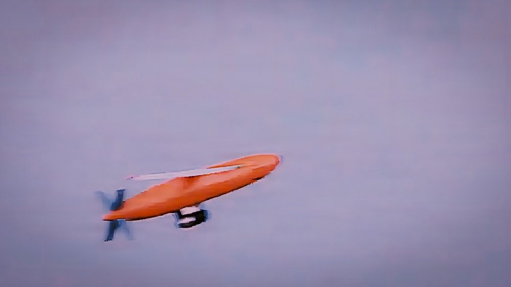 An orange fixed-wing drone or unmanned aerial vehicle (UAV) is captured in flight against an overcast, hazy sky. The aircraft has a distinctive elongated fuselage with a rear-mounted propeller and visible wing surfaces. The image appears to be a motion-blurred or slightly out-of-focus photograph taken during flight.