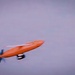 An orange fixed-wing drone or unmanned aerial vehicle (UAV) is captured in flight against an overcast, hazy sky. The aircraft has a distinctive elongated fuselage with a rear-mounted propeller and visible wing surfaces. The image appears to be a motion-blurred or slightly out-of-focus photograph taken during flight.