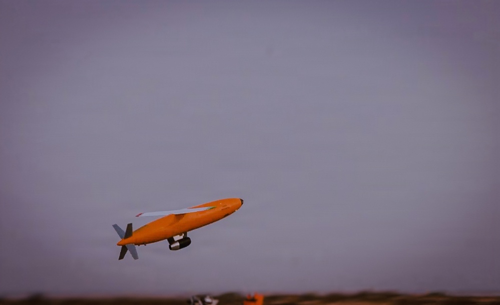 An orange unmanned aerial vehicle (UAV) or target drone is captured in flight against an overcast gray sky. The aircraft features a distinctive elongated orange fuselage with white wings and a rear-mounted propeller configuration. The image appears to be taken at dusk or dawn based on the low ambient light conditions.