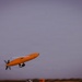 An orange unmanned aerial vehicle (UAV) or target drone is captured in flight against an overcast gray sky. The aircraft features a distinctive elongated orange fuselage with white wings and a rear-mounted propeller configuration. The image appears to be taken at dusk or dawn based on the low ambient light conditions.