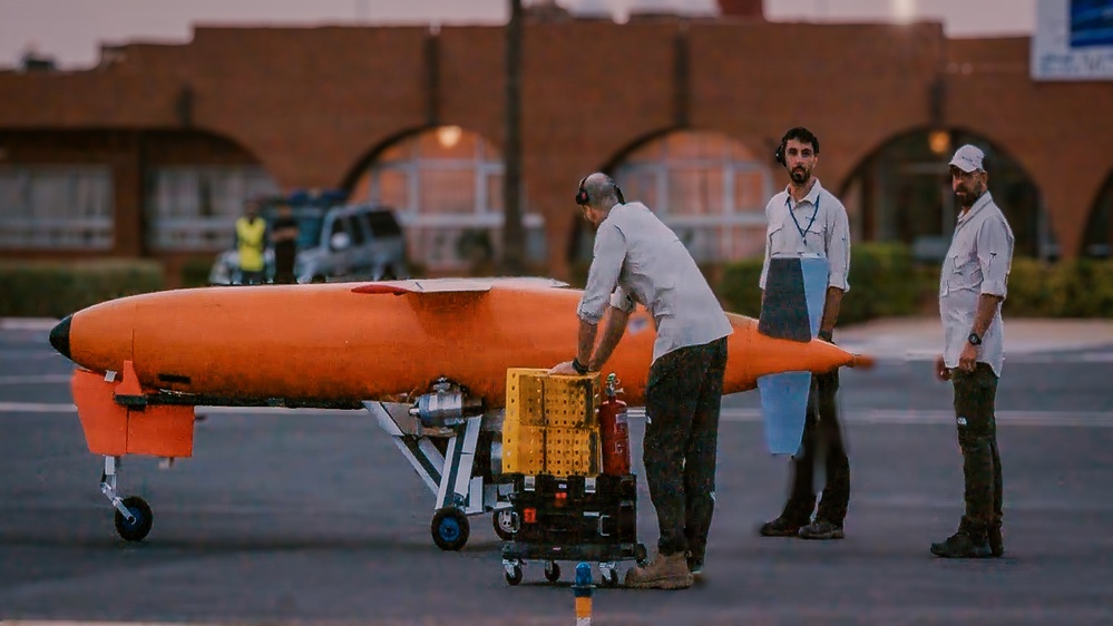 Three technicians are conducting pre-flight or maintenance operations on a bright orange aerial target drone on what appears to be an airfield tarmac at dusk. The drone is torpedo-shaped with small landing gear and is mounted on a wheeled ground support cart. A fourth person in a high-visibility vest is visible in the background near a vehicle.