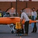 Three technicians are conducting pre-flight or maintenance operations on a bright orange aerial target drone on what appears to be an airfield tarmac at dusk. The drone is torpedo-shaped with small landing gear and is mounted on a wheeled ground support cart. A fourth person in a high-visibility vest is visible in the background near a vehicle.