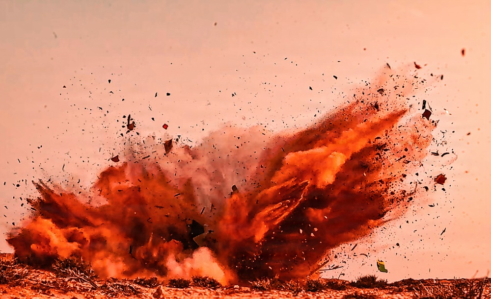 The image captures a large ground-level explosion with intense orange and red hues, likely from a controlled detonation or blast event. Debris, dust, and fragmented material are being propelled outward and upward in all directions from the blast epicenter. The explosion appears to originate at ground level in an arid or desert-like environment.