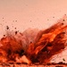 The image captures a large ground-level explosion with intense orange and red hues, likely from a controlled detonation or blast event. Debris, dust, and fragmented material are being propelled outward and upward in all directions from the blast epicenter. The explosion appears to originate at ground level in an arid or desert-like environment.