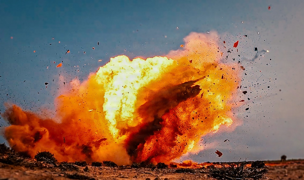 The image captures a large explosion occurring at ground level in an arid, open landscape. A massive fireball with intense yellow and orange flames is visible, accompanied by billowing smoke clouds and debris being ejected in multiple directions. The sky is clear and blue, providing strong contrast to the explosion.