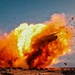 The image captures a large explosion occurring at ground level in an arid, open landscape. A massive fireball with intense yellow and orange flames is visible, accompanied by billowing smoke clouds and debris being ejected in multiple directions. The sky is clear and blue, providing strong contrast to the explosion.