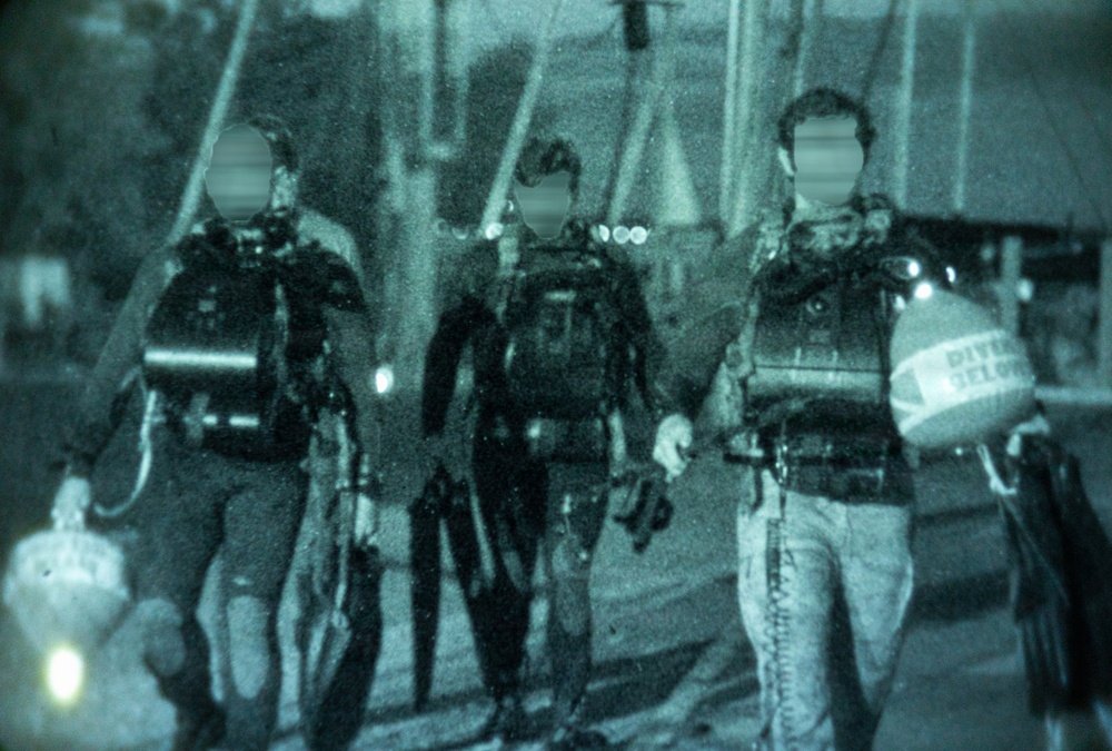 A teal-tinted archival photograph showing three individuals in full scuba diving gear walking together, likely on a dock or pier. Each person is equipped with tanks, BCDs (buoyancy control devices), and dive equipment. At least one diver is carrying a 'Diver Below' surface marker buoy.