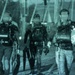 A teal-tinted archival photograph showing three individuals in full scuba diving gear walking together, likely on a dock or pier. Each person is equipped with tanks, BCDs (buoyancy control devices), and dive equipment. At least one diver is carrying a 'Diver Below' surface marker buoy.