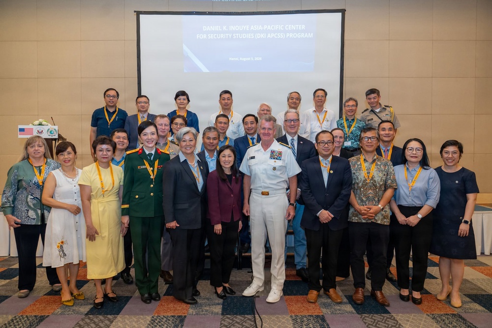 A formal group photograph taken at a Daniel K. Inouye Asia-Pacific Center for Security Studies (DKI APCSS) program event in Hanoi, dated August 5, 2026. The gathering includes military personnel, civilian officials, and program participants posing in front of a presentation screen displaying the DKI APCSS program title. The event appears to be a multilateral security studies conference or workshop with international attendees.