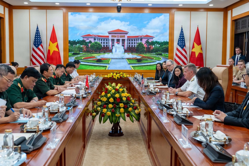 A formal bilateral meeting between Vietnamese and American military and diplomatic delegations seated at a large conference table. Both sides are represented by military officers in uniform and civilian officials, with national flags of Vietnam and the United States displayed prominently on both ends of the room. A large LED screen in the background displays an image of what appears to be a Vietnamese government building.