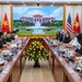 A formal bilateral meeting between Vietnamese and American military and diplomatic delegations seated at a large conference table. Both sides are represented by military officers in uniform and civilian officials, with national flags of Vietnam and the United States displayed prominently on both ends of the room. A large LED screen in the background displays an image of what appears to be a Vietnamese government building.