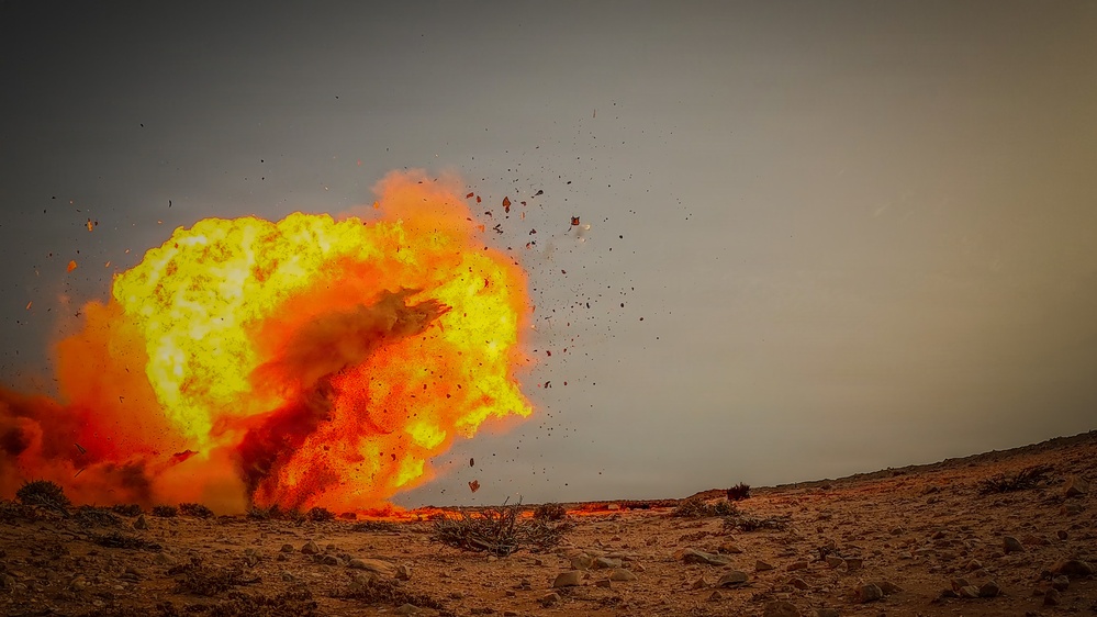 The image captures a large explosion occurring on an arid, rocky desert terrain. A prominent fireball with yellow-white core and orange-red outer flames dominates the left portion of the frame, with debris and fragments visibly ejected outward. The sky is overcast and gray, contrasting sharply with the vivid colors of the blast.