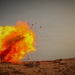 The image captures a large explosion occurring on an arid, rocky desert terrain. A prominent fireball with yellow-white core and orange-red outer flames dominates the left portion of the frame, with debris and fragments visibly ejected outward. The sky is overcast and gray, contrasting sharply with the vivid colors of the blast.