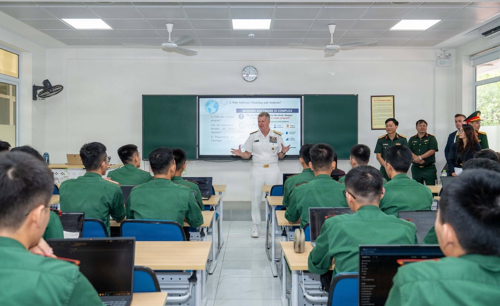 A US Navy officer in white dress uniform is delivering a lecture or presentation to a classroom of students wearing green military uniforms, likely Vietnamese military cadets. The presentation slide visible on the screen is titled 'Modern Software is Complex' under a broader topic of 'Why Software Modeling and Analysis?' Several senior military officials and civilians observe from the side of the room.