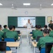 A US Navy officer in white dress uniform is delivering a lecture or presentation to a classroom of students wearing green military uniforms, likely Vietnamese military cadets. The presentation slide visible on the screen is titled 'Modern Software is Complex' under a broader topic of 'Why Software Modeling and Analysis?' Several senior military officials and civilians observe from the side of the room.
