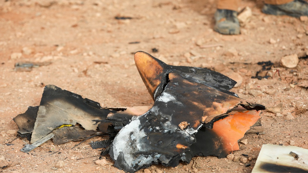 The image shows burned and charred debris lying on sandy, rocky ground. The fragments appear to be remnants of a destroyed object, showing significant fire and heat damage with blackened surfaces and melted material. A person's boot is partially visible in the upper right background.