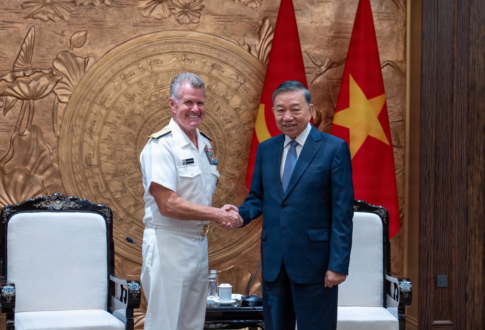 Two individuals are shaking hands in what appears to be an official diplomatic meeting. One person is wearing a white U.S. Navy dress uniform with medals and insignia, while the other is wearing a dark blue business suit. Vietnamese flags are prominently displayed in the background, indicating this is likely an official meeting in Vietnam.