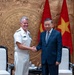 Two individuals are shaking hands in what appears to be an official diplomatic meeting. One person is wearing a white U.S. Navy dress uniform with medals and insignia, while the other is wearing a dark blue business suit. Vietnamese flags are prominently displayed in the background, indicating this is likely an official meeting in Vietnam.
