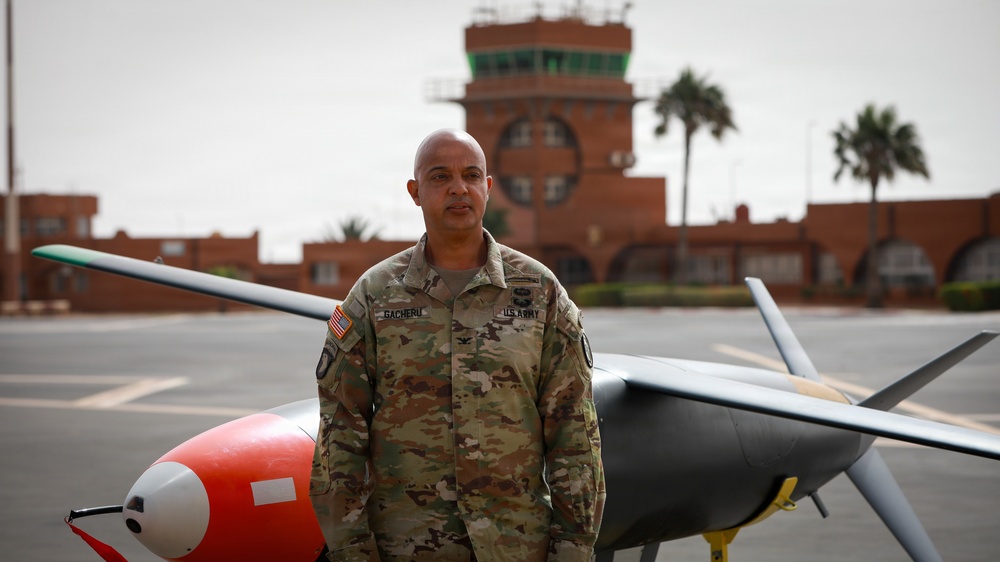A U.S. Army soldier in OCP (Operational Camouflage Pattern) uniform stands in front of a military drone/UAV on what appears to be an airfield tarmac. The soldier's name tape reads 'GACHERU' and rank insignia suggests a senior officer, with a U.S. Army patch and American flag visible. In the background, a red-brick control tower and palm trees suggest a location in a warm, possibly Middle Eastern or North African region.