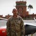 A U.S. Army soldier in OCP (Operational Camouflage Pattern) uniform stands in front of a military drone/UAV on what appears to be an airfield tarmac. The soldier's name tape reads 'GACHERU' and rank insignia suggests a senior officer, with a U.S. Army patch and American flag visible. In the background, a red-brick control tower and palm trees suggest a location in a warm, possibly Middle Eastern or North African region.