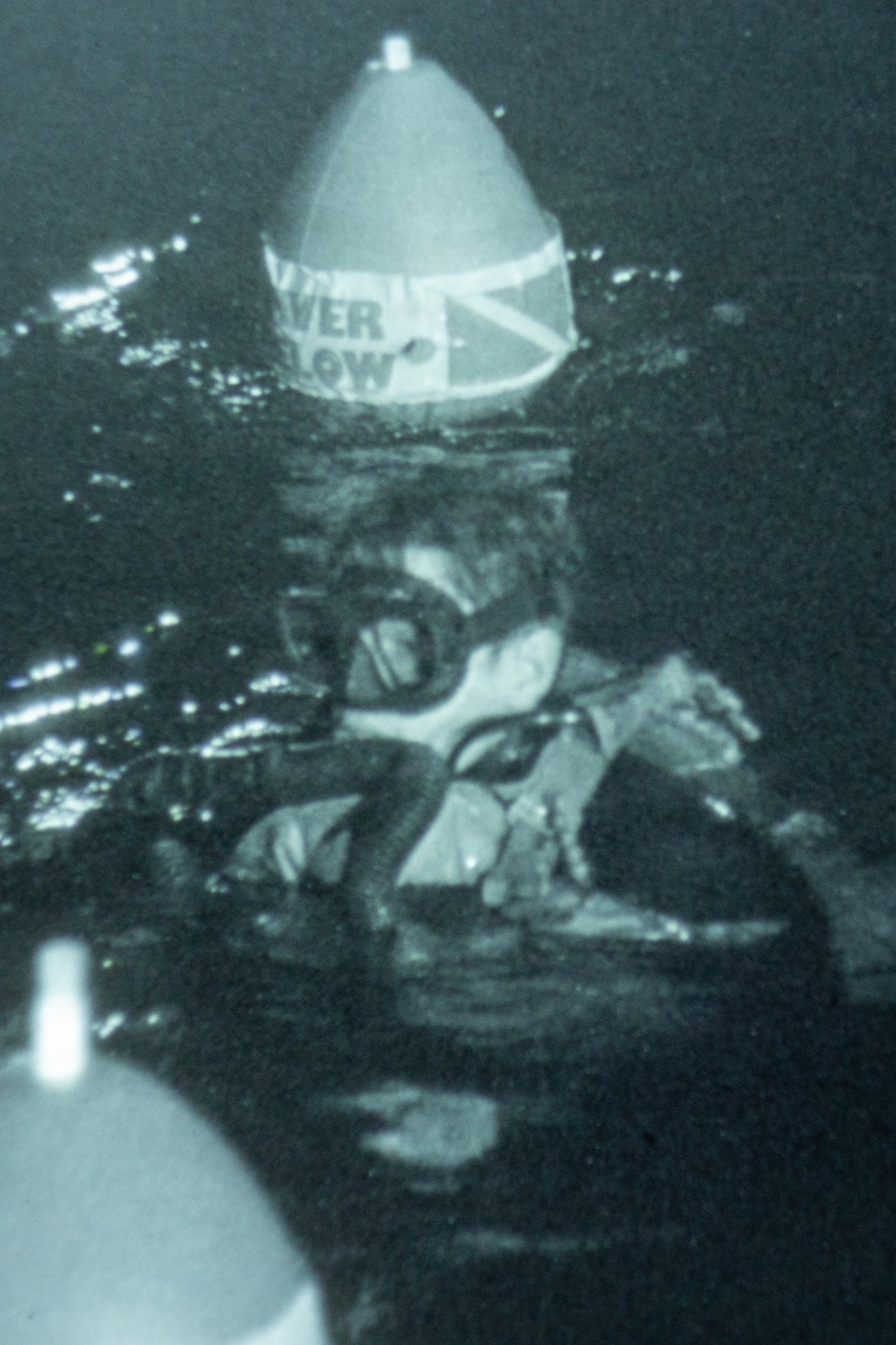 A grainy, low-quality archival photograph showing a diver underwater wearing scuba gear and diving mask. The diver appears to be positioned near or on a navigational buoy that has a conical top with partial text visible reading 'VER' and 'LOW' (likely 'OVER' and 'BELOW' or similar warning text) along with what appears to be a Union Jack (British flag) design. The image has a blue-green tint characteristic of underwater photography from an earlier era.