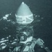 A grainy, low-quality archival photograph showing a diver underwater wearing scuba gear and diving mask. The diver appears to be positioned near or on a navigational buoy that has a conical top with partial text visible reading 'VER' and 'LOW' (likely 'OVER' and 'BELOW' or similar warning text) along with what appears to be a Union Jack (British flag) design. The image has a blue-green tint characteristic of underwater photography from an earlier era.