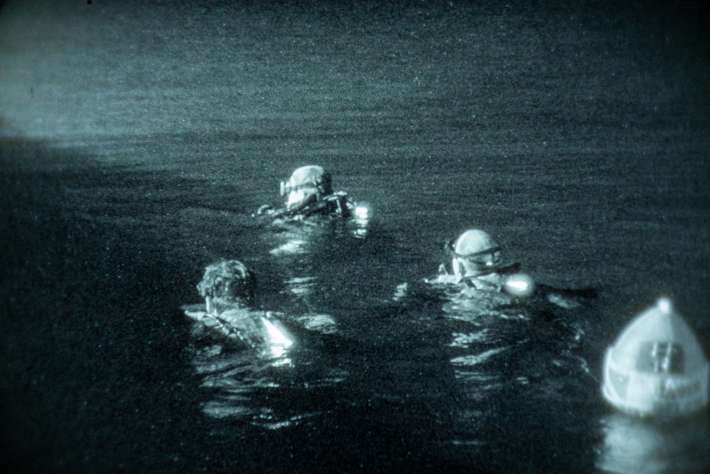 A night-vision or low-light photograph showing three individuals in the water wearing diving or tactical gear, with a small buoy or flotation device visible at the right edge. The image has a characteristic green-tinted, grainy appearance consistent with night-vision imaging technology. The scene appears to depict a nighttime water operation, possibly military or rescue in nature.