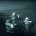 A night-vision or low-light photograph showing three individuals in the water wearing diving or tactical gear, with a small buoy or flotation device visible at the right edge. The image has a characteristic green-tinted, grainy appearance consistent with night-vision imaging technology. The scene appears to depict a nighttime water operation, possibly military or rescue in nature.