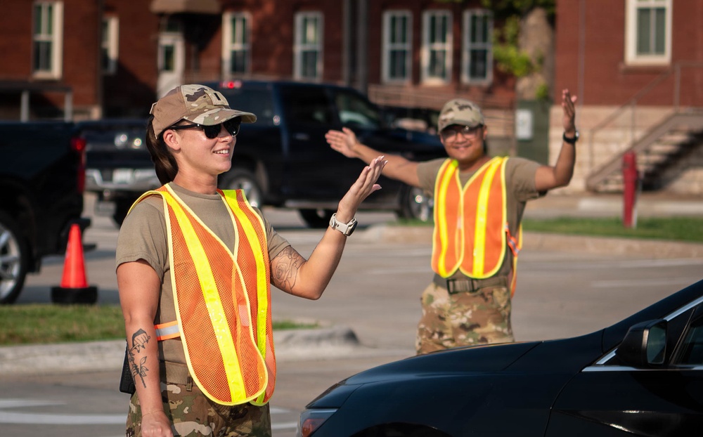 Two military personnel in camouflage uniforms and high-visibility safety vests are directing traffic in what appears to be a military installation or base. Both individuals are using hand signals to guide vehicles, with one smiling in the foreground and another visible in the background. The setting includes a brick building, parked vehicles, and orange traffic cones.