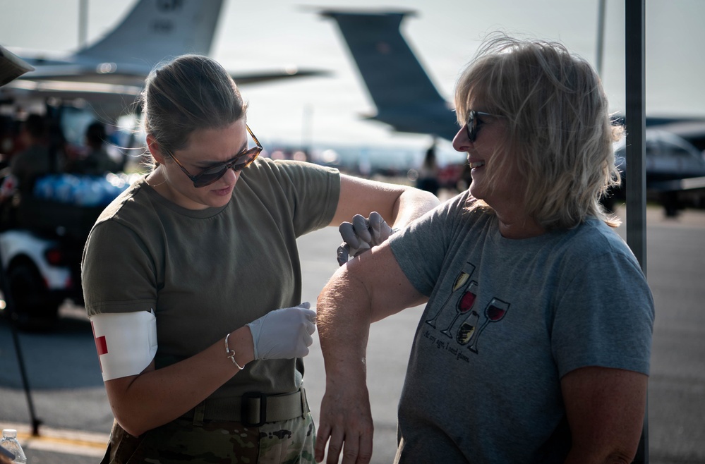 A military medical personnel wearing olive drab uniform with a red cross armband administers an injection or vaccination to a civilian woman at what appears to be an outdoor military airshow or base event. The medical provider is wearing white latex gloves and sunglasses while the civilian recipient wears a casual gray graphic t-shirt and sunglasses. Large military transport aircraft are visible in the background along with parked vehicles and other attendees.