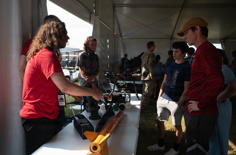 A group of people are gathered under a white tent at what appears to be an outdoor technology or aerospace exhibition. A presenter with long hair and a red shirt is demonstrating unmanned aerial vehicles (UAVs/drones) and related equipment to attendees. The setting suggests a STEM showcase, airshow, or military/defense technology event.