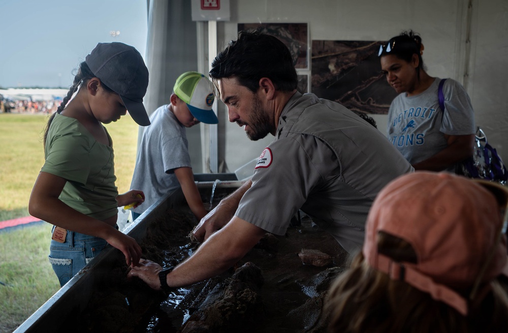 A uniformed official, likely from the U.S. Army Corps of Engineers based on the visible patch, guides children through a hands-on educational activity involving a water/sediment trough at what appears to be an outdoor public event. The interactive demonstration seems to involve examining sediment, rocks, or aquatic specimens. Several children and an adult woman in a Detroit Lions shirt observe and participate.