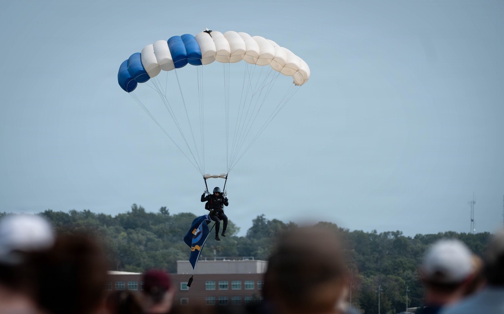 A parachutist descends toward a landing zone while carrying a flag, observed by a crowd of spectators in the foreground. The parachute is distinctly colored in blue and white panels, suggesting a military or organized demonstration team. The setting appears to be an airshow or public event, with a building and tree line visible in the background.