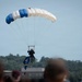 A parachutist descends toward a landing zone while carrying a flag, observed by a crowd of spectators in the foreground. The parachute is distinctly colored in blue and white panels, suggesting a military or organized demonstration team. The setting appears to be an airshow or public event, with a building and tree line visible in the background.