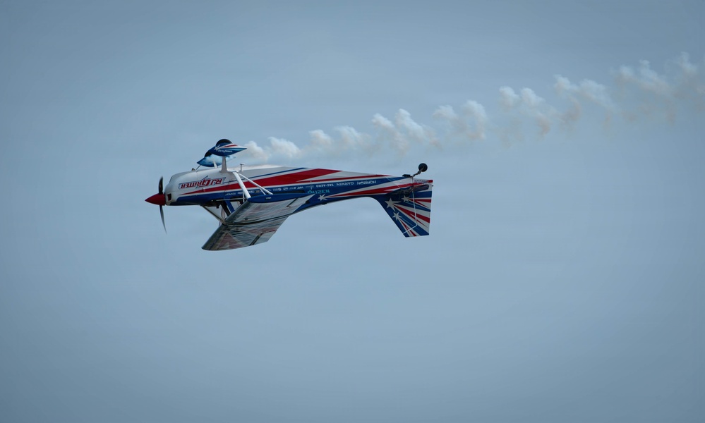 A brightly colored aerobatic aircraft is captured mid-flight performing a maneuver, appearing to be inverted or in a knife-edge position. The plane features a red, white, and blue color scheme with what appears to be an Australian flag motif on the tail. A white smoke trail streams behind the aircraft against a pale blue sky, indicating an airshow performance.