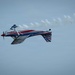 A brightly colored aerobatic aircraft is captured mid-flight performing a maneuver, appearing to be inverted or in a knife-edge position. The plane features a red, white, and blue color scheme with what appears to be an Australian flag motif on the tail. A white smoke trail streams behind the aircraft against a pale blue sky, indicating an airshow performance.