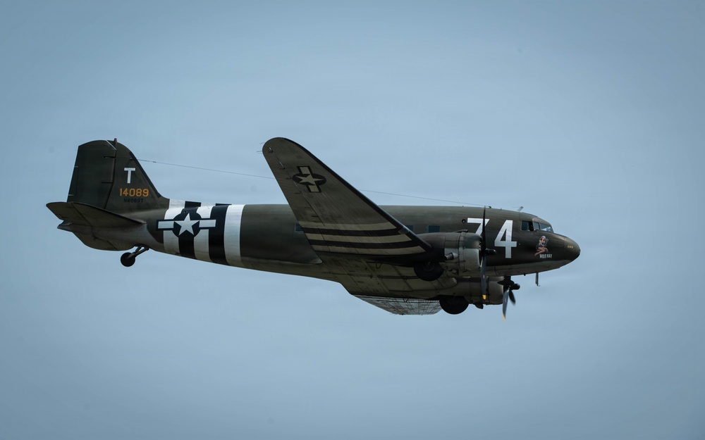 A World War II-era Douglas C-47 Skytrain military transport aircraft is captured in flight against an overcast blue-grey sky. The aircraft displays classic WWII D-Day invasion markings including black and white recognition stripes on the fuselage and wings, along with USAAF national insignia (white star in blue circle). The aircraft bears tail number 14089, the letter 'T', and the fuselage number '74', consistent with a preserved or restored warbird.