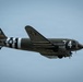 A World War II-era Douglas C-47 Skytrain military transport aircraft is captured in flight against an overcast blue-grey sky. The aircraft displays classic WWII D-Day invasion markings including black and white recognition stripes on the fuselage and wings, along with USAAF national insignia (white star in blue circle). The aircraft bears tail number 14089, the letter 'T', and the fuselage number '74', consistent with a preserved or restored warbird.